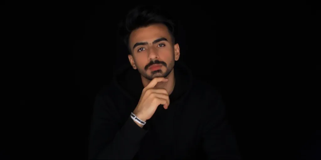 **“A young man in his twenties sitting in a completely black studio with a solid black background. He is wearing a black hoodie with a thin white wristband on his left wrist. His facial expression is neutral. The environment is dark with no neon lights. The lighting is dramatic, creating strong shadows and highlights. The image is captured from a close-up shot, focusing on his face and upper chest while clearly showing his hand. A standard focal length (50mm) is used to maintain a natural perspective.